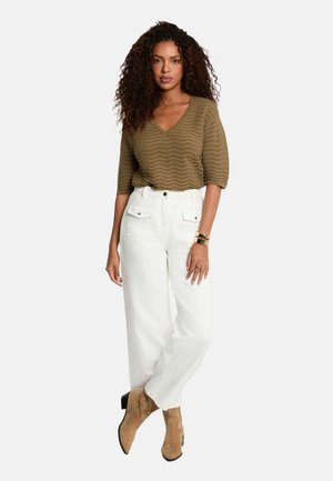 Femme aux cheveux bouclés portant un haut marron texturé à col en V, un pantalon blanc taille haute et des bottines en daim beige, debout avec une jambe croisée.