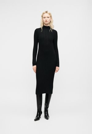 Moteris stovi dėvėdama juodą ilgarankovę, plėšytą midi suknelę su aukšta apykakle ir juodus, iki kelių siekiančius aulinukus su aukštu kulnu ant baltos fono.