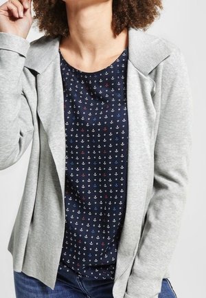 Blazer léger gris avec col replié, porté sur une chemise bleu marine à motif d'ancre répété en blanc, rouge et bleu, accompagnée de jeans slim.