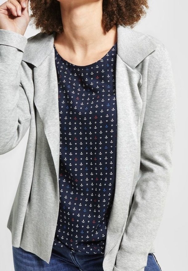 Šedý lehký blazer s ohnutým límcem, nošený přes námořnickou košili s opakujícím se vzorem kotvy v bílé, červené a modré barvě, se slim-fit džíny.