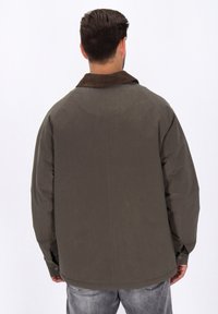 Man in een donkergroene jas met een bruine kraag, gezien vanuit de achterkant, gecombineerd met vervaagde grijze jeans.