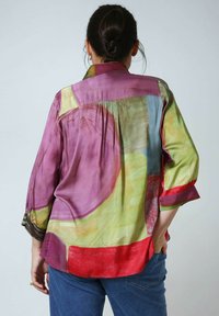 Frau mit dunklem Haar im Dutt trägt eine mehrfarbige Bluse mit abstraktem Muster und blaue Jeans, steht mit einer Hand in ihrer Tasche, mit dem Rücken zur Betrachterin.
