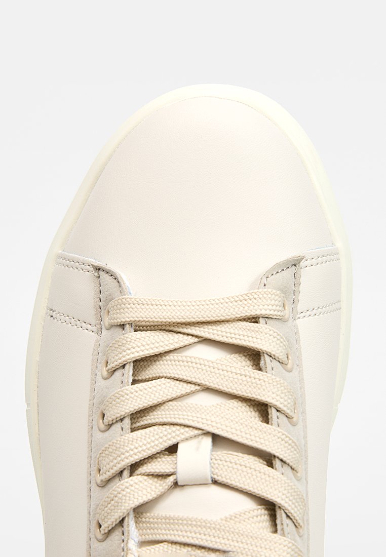 Sneaker in pelle beige con punta arrotondata, lacci piatti e un pannello laterale testurizzato. Presenta una suola in gomma leggera a contrasto.