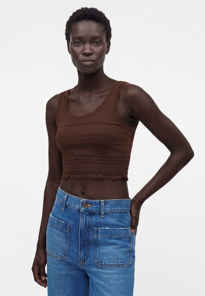 Γυναίκα με κοντά μαλλιά που φοράει ένα αμάνικο καφέ crop top με υφασμάτινες λωρίδες και μπλε ψηλόμεσα τζιν με μεγάλες μπροστινές τσέπες