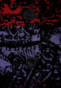 Tessuto decorato in rosso, viola e nero. Presenta un prominente bottone nero con testo in rilievo. La texture appare liscia con un design intricato.