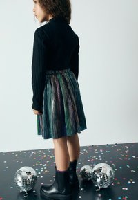 Top a maniche lunghe nero a costine abbinato a una gonna plissettata multicolore. L'outfit è completato da stivaletti neri e coriandoli sparsi sul pavimento.