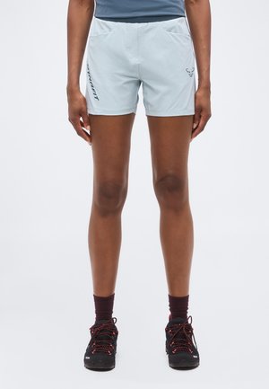 Osoba nosící světle modré sportovní kraťasy, tmavé ponožky a černé sportovní boty, stojící vzpřímeně před jednoduchým pozadím.