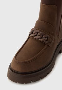 Bruine leren enkellaars met een chunky zool, voorzien van een decoratieve kettingaccent en een gladde textuur. Ontworpen voor comfort en stabiliteit.