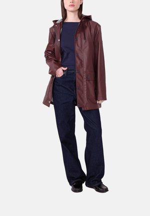 Frau trägt eine braune Kapuzenregenjacke, dunkelblaues Oberteil, dunkelblaue Jeans und schwarze Schuhe, steht mit einer Hand in der Tasche.