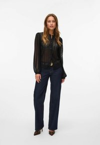 Must mustrõnglace blouse, millel on nööpidega ees, pikad varrukad ja avatud kudumus, koos tumedate siniste laiad jalad teksade ja kontsadega kingadega.