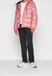 Różowa kurtka puchowa z kapturem, zapinana na zamek, z bocznymi kieszeniami, w zestawieniu z czarnymi spodniami i białymi trampkami z pomarańczowymi akcentami.