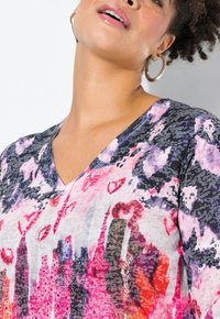 V-ringad tunika i flerfärgad tyg med abstrakta mönster, paljetter och en mjuk textur. Designen domineras av främst rosa och lila toner.