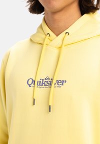 Sudadera con capucha amarilla, fabricada en un tejido suave, que cuenta con cordones y un logo en la parte delantera. La textura parece lisa, sin patrones visibles.