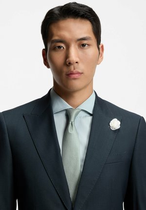 Completo scuro su misura con rever affilato, camicia azzurro chiaro e cravatta verde chiaro con motivo. Accento floreale bianco sul rever. Tessuto liscio.