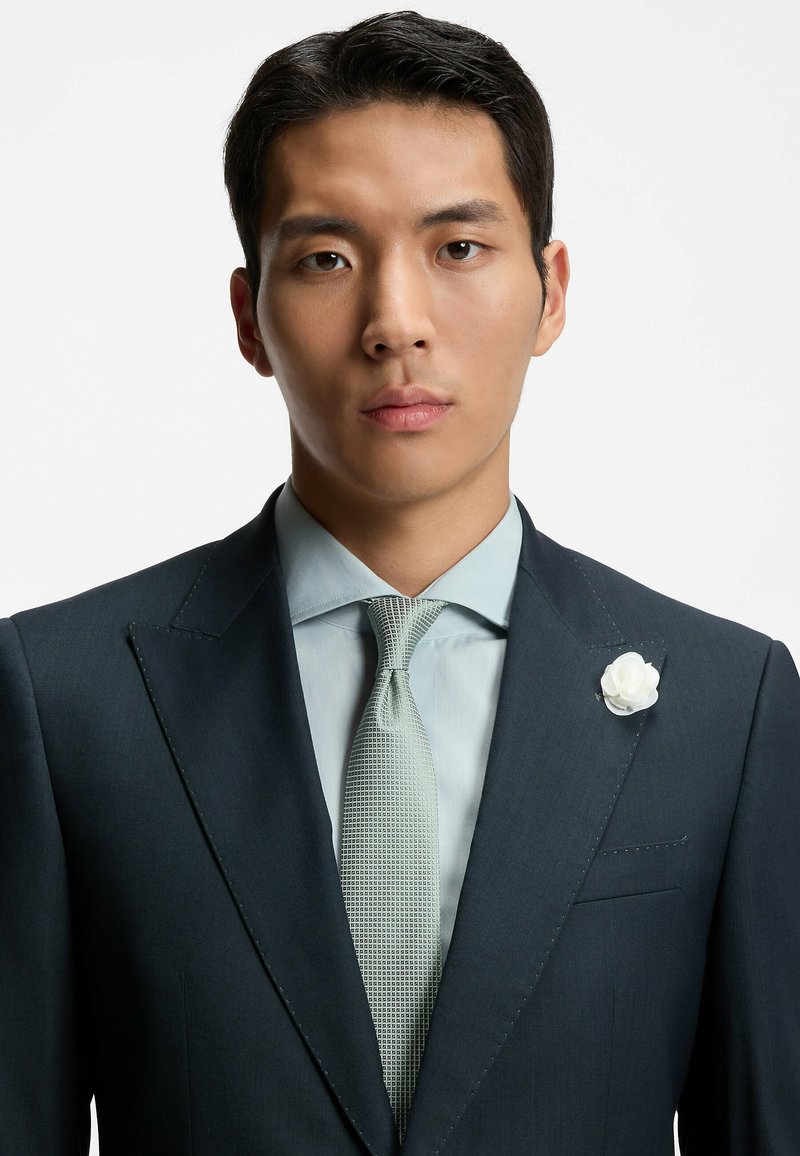 Costume sombre ajusté avec un revers marqué, chemise bleu clair et cravate verte claire à motif. Accent de fleur blanche sur le revers. Texture lisse.