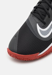 Černá sportovní obuv s texturovaným svrškem, šedými akcenty a červenou gumovou podrážkou. Obsahuje výrazný bílý znak Nike.