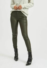 Olivi vihreät keinonahkalegginsit, joissa on slim fit -tyyli ja sivusaumat. Yhdistetty vaaleanharmaaseen neuleeseen ja mustiin nilkkasaappaisiin.