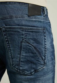 Jeans de denim con un lavado de azul oscuro, costuras en contraste, un bolsillo de parche con diseño asimétrico y una etiqueta de cuero negro en la cintura.