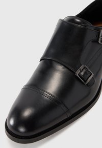 Scarpa in pelle nera con punta arrotondata, accentata da doppia fibbia e dettagli cuciti. Texture liscia; design elegante e moderno.