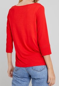 Haut rouge à manches trois-quarts avec un large col et des coutures décoratives le long de l'ourlet des manches. Assorti avec un jean en denim bleu.
