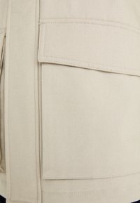 Giacca in tela beige con ampie tasche a patta, cuciture piatte e design strutturato. Il tessuto ha una texture liscia e resistente.