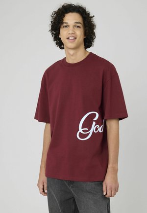 T-shirt en coton bordeaux avec une coupe décontractée, présentant un grand graphisme blanc "Good" sur le devant. Manches courtes et design col rond.