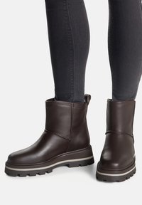 Tmavohnedé kožené členkové čižmy s hrubou podrážkou a béžovým pruhom, nosené s priliehavými čiernymi nohavicami, viditeľnými od polovice holene nadol.