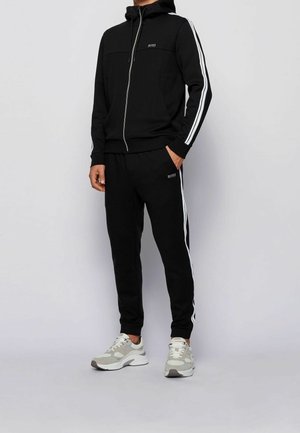 Survêtement noir en mélange de coton ; hoodie zippé avec poches latérales, rayures blanches sur les manches et le pantalon, agrémenté d'un logo. Baskets grises.