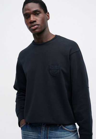 Νεαρός άνδρας που φοράει ένα μαύρο φούτερ με μακριά μανίκια και ένα μικρό κομμένο λογότυπο στο αριστερό στήθος και μπλε τζιν, στέκεται μπροστά σε λευκό φόντο.