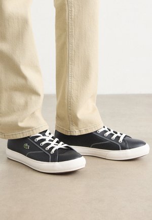 Person iført beige bukser og sorte lærreds-sneakers med hvide såler og snørebånd, stående på en glat beige gulv.