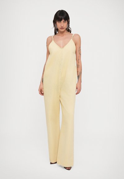 Γυναίκα με σκούρα μαλλιά που φοράει μια ανοιχτόκκινη, κίτρινη αμάνικη ολόσωμη φόρμα με φαρδιά πόδια και σανδάλια, στέκεται μπροστά σε λευκό φόντο.