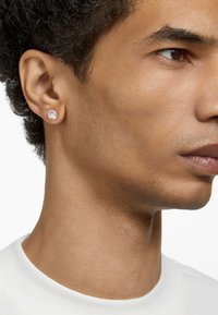 Kulaté náušnice se středovým průhledným kamínkem, obklopené kovovým rámem ve růžovém zlatě. Modelka má náušnici na pozadí světlé pleti.