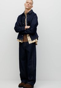 Temno denim jakna in široke hlače z ravnimi nogami, kombinirane s rjavo srajco in karo flanelasto majico. Rjavi čevlji z gladko teksturo in minimalnimi kovinskimi detajli.