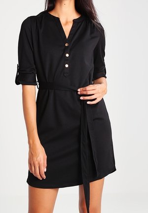 Nainen, joka pukeutunut mustaan napilliseen mekkoon, jonka hihat on käännetty ja jossa on vyötärönauha, seisoo yksinkertaista valkoista taustaa vasten.