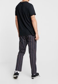 T-shirt en coton noir associé à un pantalon à rayures sombres avec des lignes rouges et blanches, complété par des baskets noires et des chaussettes blanches mi-mollet.