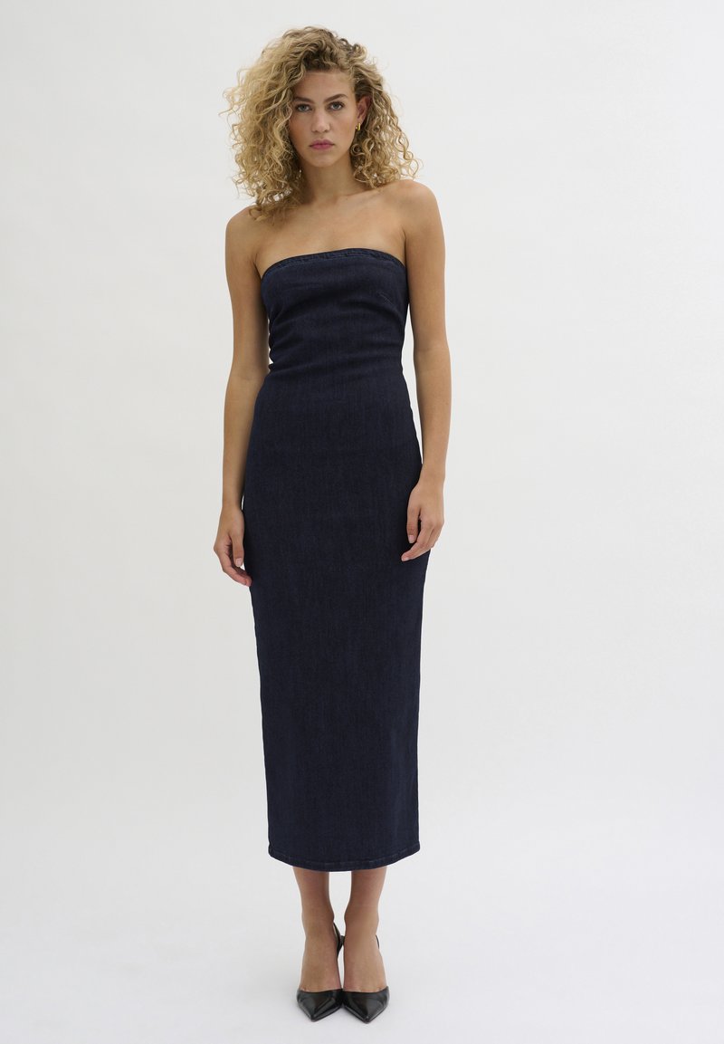 Abito in denim fitted senza spalline, di colore blu scuro, fino al ginocchio; presenta una silhouette dritta e una texture liscia, indossato con tacchi neri a punta.