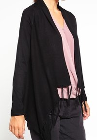 Svart sjal med fransar och öppen framsida, tillverkad av ett mjukt material. Den har en draperad design och är kombinerad med en ljusrosa topp under.