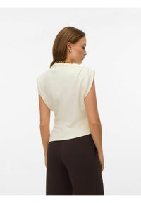 Blusa senza maniche color crema con drappeggio sulle spalle, design aderente e dettaglio con cucitura posteriore, abbinata a pantaloni marrone scuro e una collana di perline.