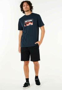Camiseta de algodón azul marino con un colorido logo de RIP CURL, combinada con pantalones cortos negros. Las zapatillas son negras con detalles blancos, llevadas con calcetines negros.