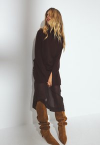 Un gros pull marron par-dessus une jupe noire transparente, associé à des bottes en daim moutarde, hautes et décontractées. Couleurs unies, textures lisses.