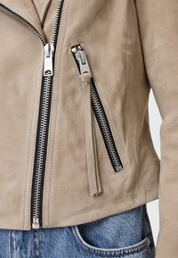 Beige suède motorjack met een diagonale rits, zwarte accenten en twee zijzakken, met een soepele textuur en zilverkleurige hardware.