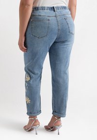 Ljusblå denimjeans med elastisk midja, uppvikta fållar och blommönster broderat på vänster ben. Bakfickor synliga.