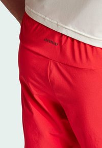 Un short de sport rouge présente une taille élastique avec le logo "AEROREADY". Le matériau semble lisse, léger et respirant.