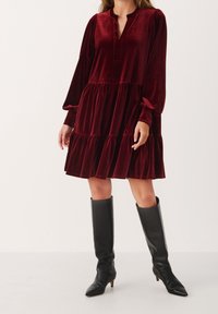 Burgundivärinen samettimekko, jossa on pitkät hihat, V-muotoinen kaula-aukko ja kerroksellinen hame. Yhdistetty polvipitkiin mustiin nahkabootseihin. Suura tekstuuri.