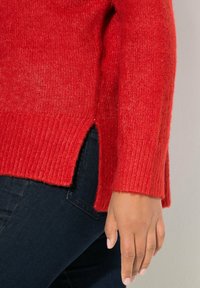 Roter Strickpullover mit geripptem Muster, seitlichen Schlitzen und lockerer Passform. Kombiniert mit dunkelblauen Jeans und einem sanften, neutralen Hintergrund.