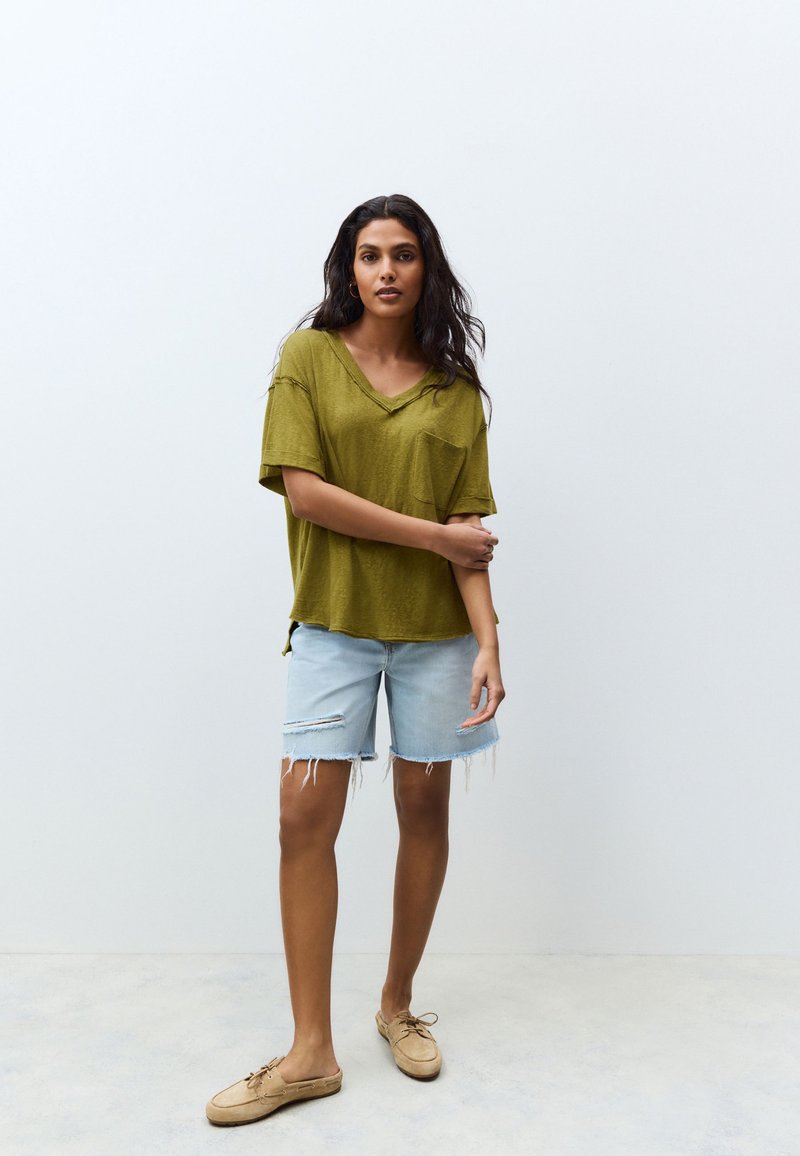 Olivengroen V-hals T-shirt met een voorzak, gecombineerd met lichtblauwe denim shorts met een gescheurde zoom en rafelige randen, gedragen met beige schoenen.