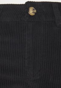 Zwarte corduroy stof met verticale ribbels, voorzien van een knoopsluiting en een klassiek ontwerp van de tailleband. Details omvatten een klein zakje.