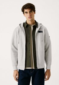 Sudadera gris con cremallera y capucha ajustable, puños acanalados y un bolsillo en el pecho, llevada sobre una camiseta verde oscura y una blusa clara.