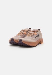 Chaussures de trail beige clair avec tige en maille texturée, accents orange et semelle violette robuste pour une meilleure traction.