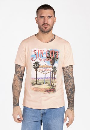Uomo con tatuaggi indossa una maglietta color pesca con un design "Sin City Las Vegas" con palme e paesaggio desertico.